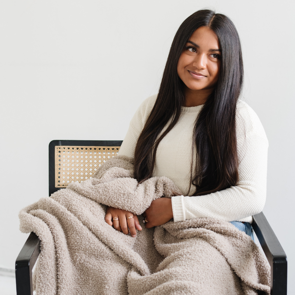 A woman sits with a Bamboni® blanket by Saranoni. This ultra-soft blanket is a taupe blanket and made from a premium blend of bamboo rayon and poly microfiber, offering a lightweight, breathable, and fluffy feel. Perfect as a extra large blanket, luxury blanket, plush blanket, or soft blanket, it adds warmth and elegance to any living room, bedroom, or home décor setting. Designed for ultimate comfort, this buttery-soft Saranoni blanket is ideal for gifting. Enjoy this cozy blanket for years to come! 