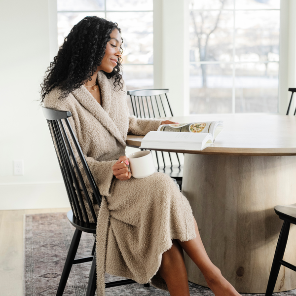 A woman reads while wearing a Saranoni Bamboni® Robe in Taupe. Crafted from our signature ultra-soft and breathable Bamboni® fabric, this women’s plush robe wraps you in warmth while remaining lightweight. Featuring a classic shawl collar, deep pockets, and an adjustable tie waist, it's perfect for lounging, self-care mornings, and unwinding. Elevate your relaxation with the best soft robe for women, designed for everyday luxury. The soft robe is a luxury robe and a Saranoni robe. 