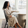 A woman is sitting at a table and wearing a Saranoni Bamboni® Robe in Taupe. Crafted from our signature ultra-soft and breathable Bamboni® fabric, this women’s plush robe wraps you in warmth while remaining lightweight. Featuring a classic shawl collar, deep pockets, and an adjustable tie waist, it's perfect for lounging, self-care mornings, and unwinding. Elevate your relaxation with the best soft robe for women, designed for everyday luxury. The soft robe is a luxury robe and a Saranoni robe. 