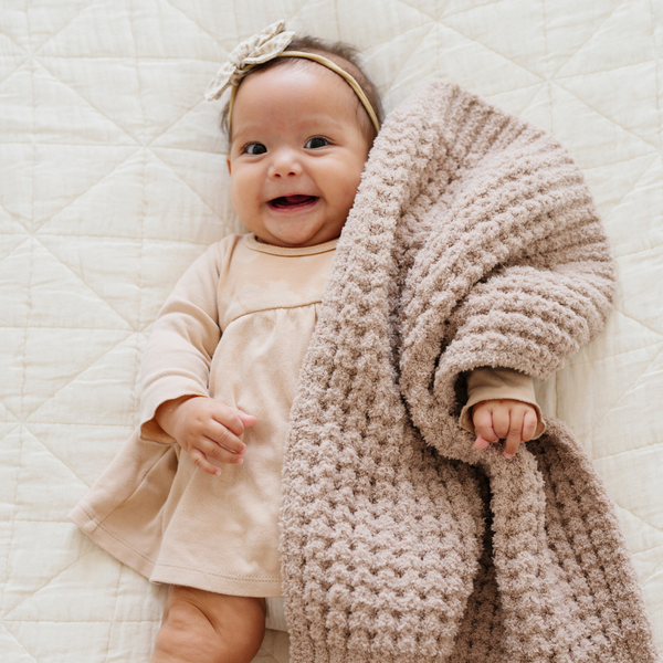 Happy baby in a neutral outfit lying on a quilted cream blanket, wrapped in a soft tan blanket that is a waffle knit blanket as well as a mini blanket and baby blanket by Saranoni. This cozy, textured infant blanket in warm beige is perfect for snuggles and comfort. This is a cozy Saranoni blanket. 