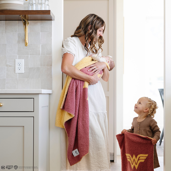 A mother holds her baby girl in a pink and yellow double-layer Bamboni® receiving blanket with a Wonder Woman™ icon on it while standing by her other daughter who is holding the same blanket is a mini size. The soft blankets are luxury blankets and Saranoni blankets. These kids blankets are perfect for littles. 