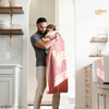 A father holds his daughter in a pink and yellow double-layer Bamboni® blanket with a Wonder Woman™ icon on it. The soft blanket is a luxury blanket and a receiving blanket as well as a Saranoni blanket. This kids blanket is perfect for littles. 