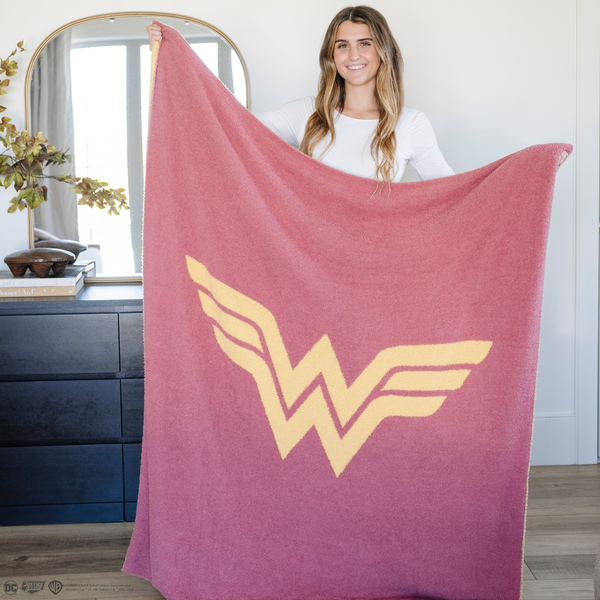 A woman holds a pink and yellow double-layer Bamboni® blanket with a Wonder Woman™ icon on it. The soft blanket is a luxury blanket and a throw blanket as well as a Saranoni blanket.