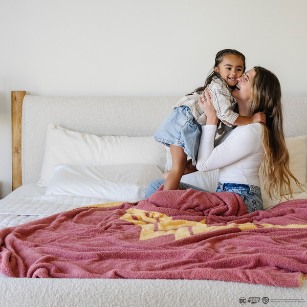 A mother holds her daughter. They are by a pink and yellow double-layer Bamboni® blanket with a Wonder Woman™ icon on it. The soft blanket is a luxury blanket and a throw blanket as well as a Saranoni blanket. 