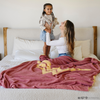 A mother holds her daughter. They are by a pink and yellow double-layer Bamboni® blanket with a Wonder Woman™ icon on it. The soft blanket is a luxury blanket and a throw blanket as well as a Saranoni blanket. 