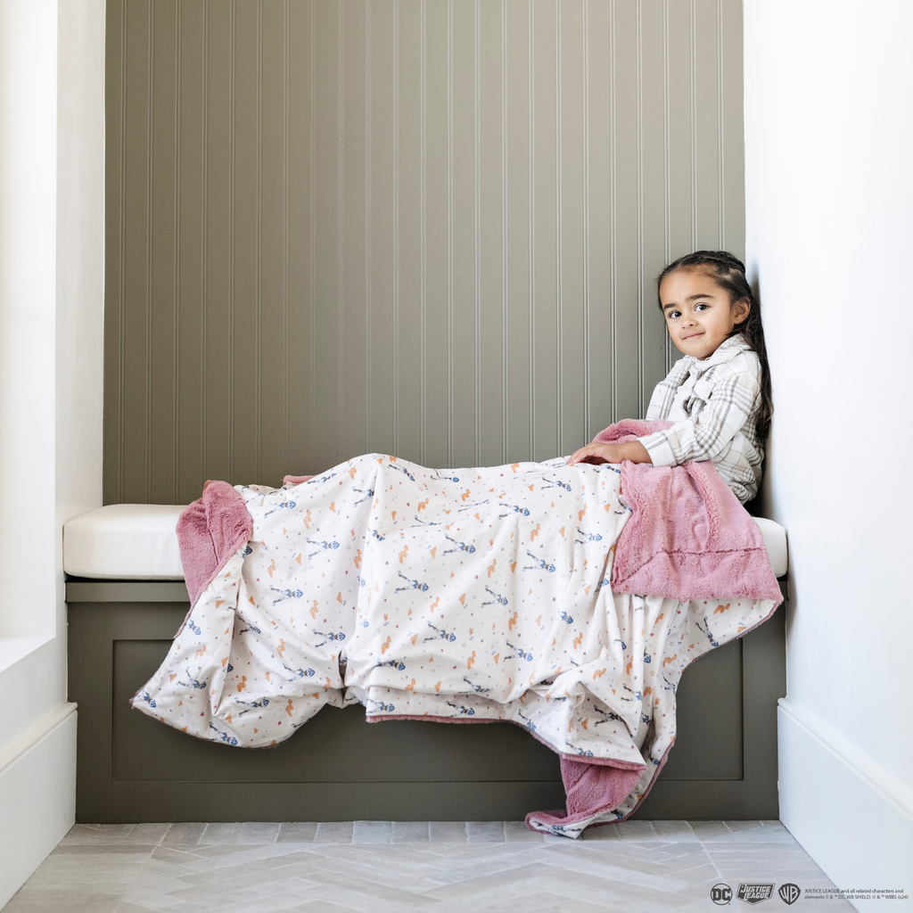 A little girl sits under a soft minky blanket showcasing Wonder Woman™ and various Wonder Woman™ icons. The white background enhances the vibrant details, while the pink/coral plush underside adds extra coziness. Ideal for snuggling, showcasing superhero pride, or adding a fun, adventurous touch to your decor. The soft blanket is a Saranoni blanket. 