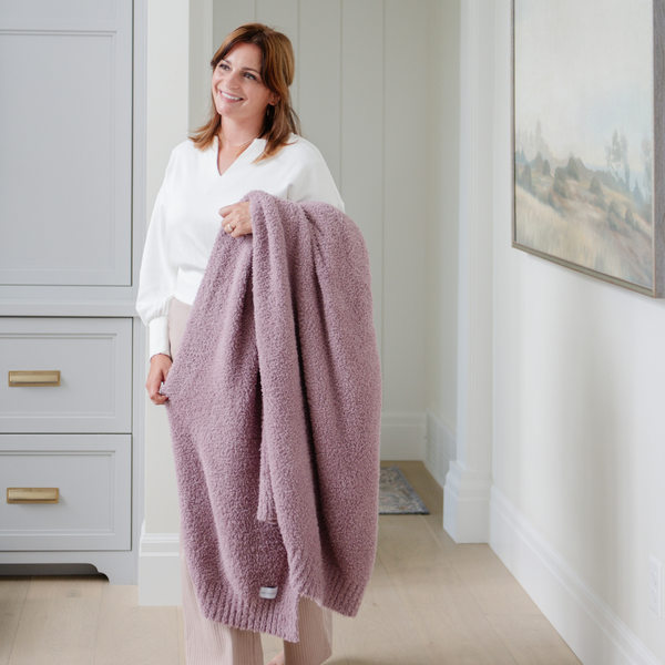 A woman smiles and carries a Bamboni® blanket by Saranoni. This ultra-soft blanket is a light purple blanket and made from a premium blend of bamboo rayon and poly microfiber, offering a lightweight, breathable, and fluffy feel. Perfect as a extra large blanket, luxury blanket, or soft blanket, it adds warmth and elegance to any living room, bedroom, or home décor setting. Designed for ultimate comfort, this buttery-soft Saranoni blanket is ideal for gifting. Enjoy this cozy blanket for years to come!