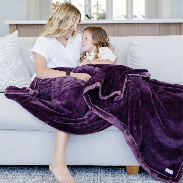 A mother holds her little girl while sitting under a Luxury Extra Large Oversized Throw Aubergine (deep purple) Colored Lush Saranoni Blanket. The soft blanket is an extra large throw blanket and has a soft faux fur feeling.