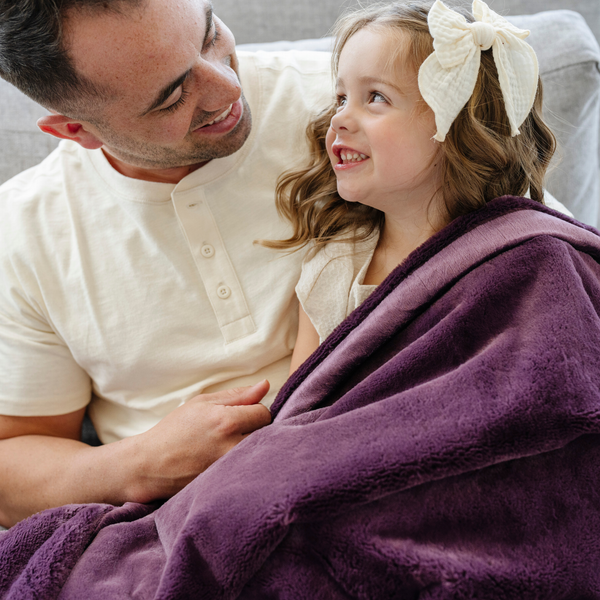 A father holds his little girl while sitting under a Luxury Extra Large Oversized Throw Aubergine (deep purple) Colored Lush Saranoni Blanket. The soft blanket is an extra large throw blanket and has a soft faux fur feeling.