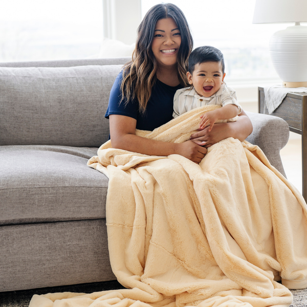 A mother and her little boy smile and sit under a Chiffon (light yellow) Lush XL Saranoni blanket, designed for oversized comfort and featuring a buttery soft, plush texture. This luxurious Saranoni blanket is perfect for cozy evenings, reading, or home decor. The Saranoni blanket adds warmth, style, and unmatched softness, making it an ideal gift or essential for ultimate relaxation.