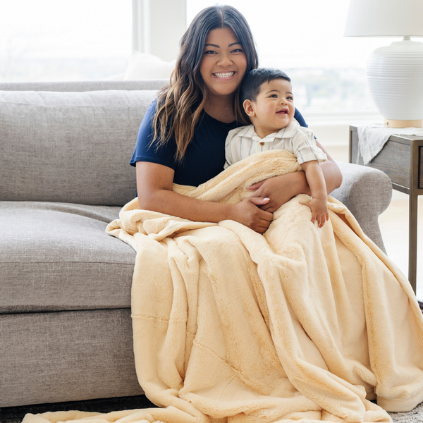 A woman holds her little boy while sitting under a Luxury Extra Large Oversized Throw Chiffon (light yellow) Colored Lush Saranoni Blanket. The soft blanket is an extra large throw blanket and has a soft faux fur feeling.