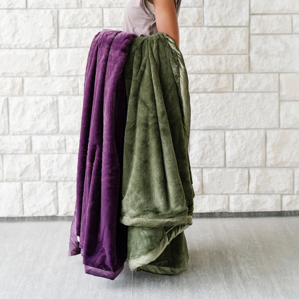 A woman holds a Luxury Extra Large Oversized Throw Aubergine (deep purple) Colored Lush Saranoni Blanket and a Luxury Extra Large Oversized Throw Fern Colored Lush Saranoni Blanket. The soft blanket is an extra large throw blanket and has a soft faux fur feeling.