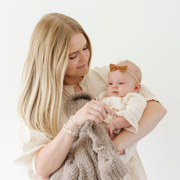 A mother holds her little girl in a Taupe colored Bamboni® Saranoni Blanket. The soft blanket is a small blanket and a baby blanket as well as a mini blanket.