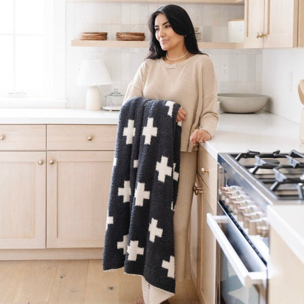 A woman holds a luxurious double-layer Bamboni® blanket by Saranoni, featuring a charcoal and white blanket with a swiss cross pattern. This ultra-soft blanket is crafted from bamboo rayon and poly microfiber, making it lightweight, breathable, and fluffy. Ideal as a cozy throw blanket, luxury blanket, plush throw, soft blanket, or stylish home accent, this Saranoni blanket brings warmth, comfort, and timeless elegance to any living room or bedroom.