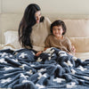 A mother and her son smile and sit with a luxurious double-layer Bamboni® blanket by Saranoni, featuring a charcoal and white blanket with a swiss cross pattern. This ultra-soft blanket is crafted from bamboo rayon and poly microfiber, making it lightweight, breathable, and fluffy. Ideal as a cozy blanket, extra large blanket, luxury blanket, soft blanket, or stylish home accent, this Saranoni blanket brings warmth, comfort, and timeless elegance to any living room or bedroom.