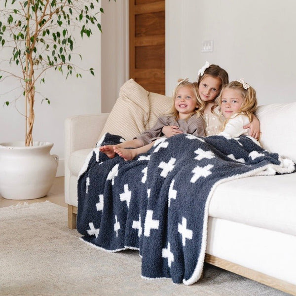 Three little girls sit with a luxurious double-layer Bamboni® blanket by Saranoni, featuring a charcoal and white blanket with a swiss cross pattern. This ultra-soft blanket is crafted from bamboo rayon and poly microfiber, making it lightweight, breathable, and fluffy. Ideal as a cozy blanket, extra large blanket, luxury blanket, soft blanket, or stylish home accent, this Saranoni blanket brings warmth, comfort, and timeless elegance to any living room or bedroom.