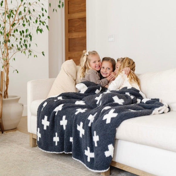 Three little girls hug and sit with a luxurious double-layer Bamboni® blanket by Saranoni, featuring a charcoal and white blanket with a swiss cross pattern. This ultra-soft blanket is crafted from bamboo rayon and poly microfiber, making it lightweight, breathable, and fluffy. Ideal as a cozy blanket, extra large blanket, luxury blanket, soft blanket, or stylish home accent, this Saranoni blanket brings warmth, comfort, and timeless elegance to any living room or bedroom.