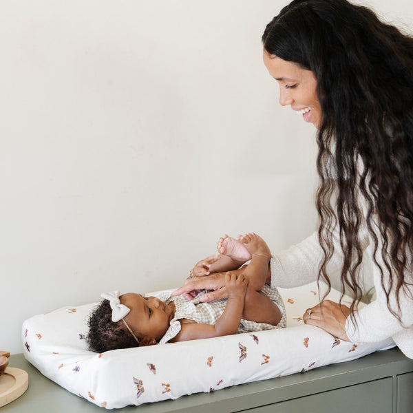 A mother playing her little girl who is laying on an ultra-soft Saranoni Minky Stretch Luxe changing cover in a butterfly pattern, crafted from 94% polyester and 6% spandex for extra stretch and durability. Perfect for any baby nursery, this breathable, stretchy changing pad cover ensures a snug fit while adding a touch of style. Ideal for parents seeking high-quality baby essentials that are soft, luxurious, and easy to care for – wash cold, no softeners, hang or lay flat to dry.