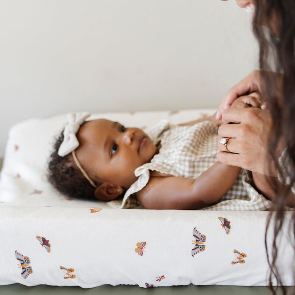 A mother playing her little girl who is laying on an ultra-soft Saranoni Minky Stretch Luxe changing cover in a butterfly pattern, crafted from 94% polyester and 6% spandex for extra stretch and durability. Perfect for any baby nursery, this breathable, stretchy changing pad cover ensures a snug fit while adding a touch of style. Ideal for parents seeking high-quality baby essentials that are soft, luxurious, and easy to care for – wash cold, no softeners, hang or lay flat to dry.