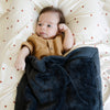 A baby boy lays on a Minky Stretch Luxe swaddle in a star pattern, featuring ultra-soft minky fabric with a luxurious stretch, perfect for keeping your baby cozy and comfortable. Ideal for newborns and infants. He as a charcoal lush Saranoni baby blanket as well.