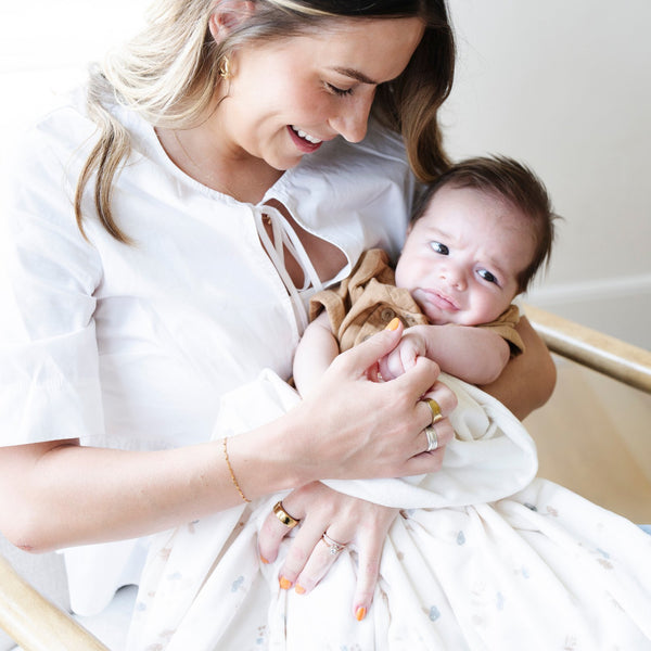 A mother holds her baby boy in a Minky Stretch Luxe swaddle in a mushroom pattern, featuring ultra-soft minky fabric with a luxurious stretch, perfect for keeping your baby cozy and comfortable. Ideal for newborns and infants.