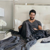 A man sits while holding a Luxury Extra Large Oversized Throw Charcoal Colored Lush Saranoni Blanket. The soft blanket is an extra large throw blanket and has a soft faux fur feeling.