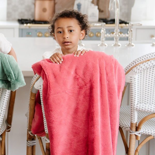 A little girl sits with a Luxury Receiving Primrose (bold pink) Colored Lush Saranoni Blanket. The soft blanket is a small blanket and a baby blanket or toddler blanket.