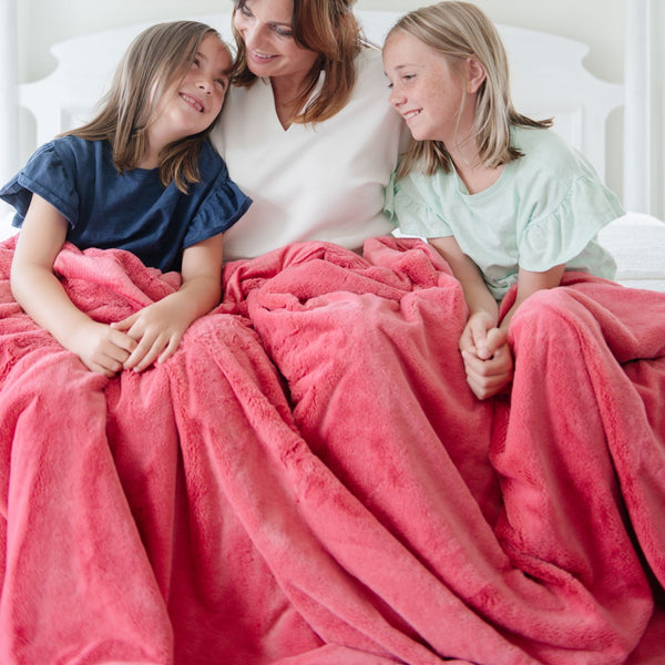 A mother and her two daughters sit under a Luxury Extra Large Oversized Throw Primrose (bold pink) Colored Lush Saranoni Blanket. The soft blanket is an extra large throw blanket and has a soft faux fur feeling.