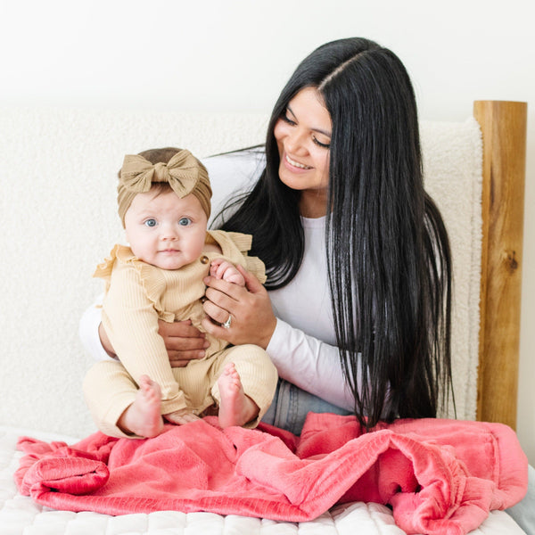 A mother holds her baby girl over a Luxury Receiving Primrose (bold pink) Colored Lush Saranoni Blanket. The soft blanket is a small blanket and a baby blanket or toddler blanket.