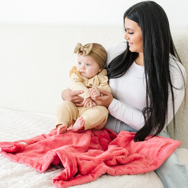 A mother holds her baby girl on a Luxury Receiving Primrose (bold pink) Colored Lush Saranoni Blanket. The soft blanket is a small blanket and a baby blanket or toddler blanket.