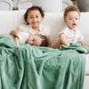 A little girl and her two brothers sit holding a Toddler Seagrass (dusty green) Colored Lush Luxury Saranoni Blanket. The soft blanket is a baby blanket or toddler blanket.