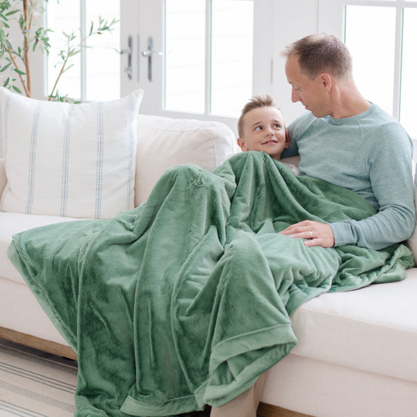 A father and his son sit under a Luxury Extra Large Oversized Throw Seagrass (dusty green) Colored Lush Saranoni Blanket. The soft blanket is an extra large throw blanket and has a soft faux fur feeling.