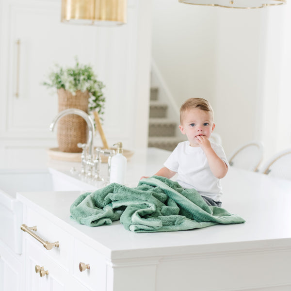 A little boy sits under a Luxury Receiving Seagrass (dusty green) Colored Lush Saranoni Blanket. The soft blanket is a small blanket and a baby blanket or toddler blanket.