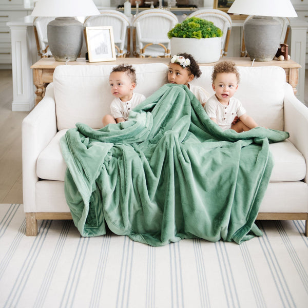 A little girl and her two brothers sit under a Luxury Extra Large Oversized Throw Seagrass (dusty green) Colored Lush Saranoni Blanket. The soft blanket is an extra large throw blanket and has a soft faux fur feeling.