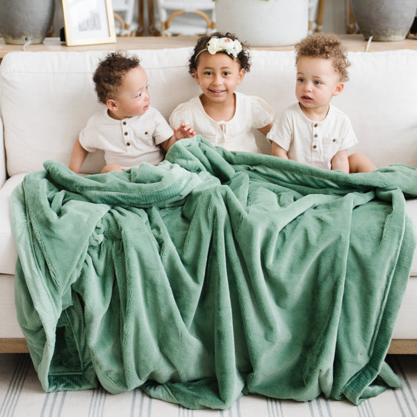 A little girl and her two brothers sit under a Luxury Extra Large Oversized Throw Seagrass (dusty green) Colored Lush Saranoni Blanket. The soft blanket is an extra large throw blanket and has a soft faux fur feeling.