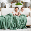 A little girl and her two brothers sit under a Luxury Extra Large Oversized Throw Seagrass (dusty green) Colored Lush Saranoni Blanket. The soft blanket is an extra large throw blanket and has a soft faux fur feeling.