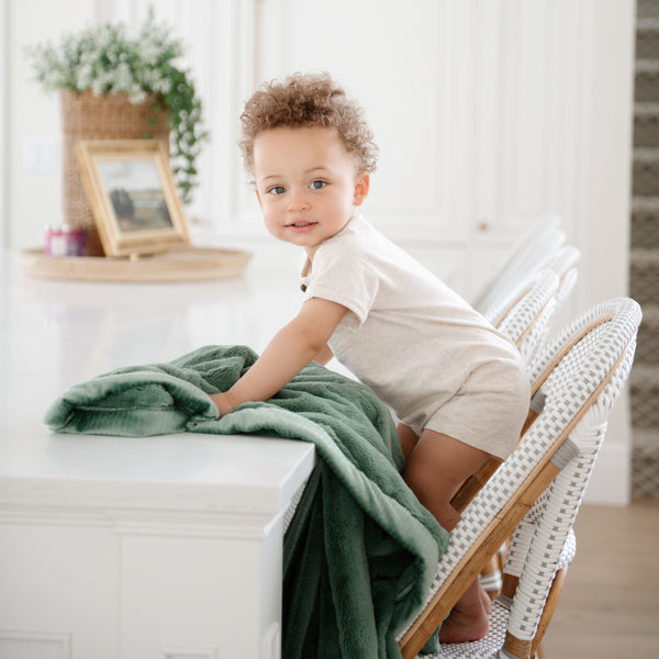 A little boy stands with a Luxury Receiving Seagrass (dusty green) Colored Lush Saranoni Blanket. The soft blanket is a small blanket and a baby blanket or toddler blanket.