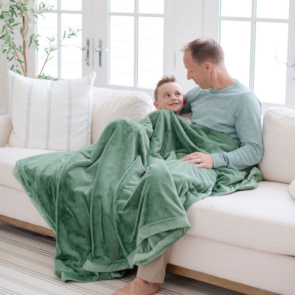 A father and son sit under a Seagrass (dusty green) Colored Lush Luxury Saranoni Blanket. It is a soft blanket.