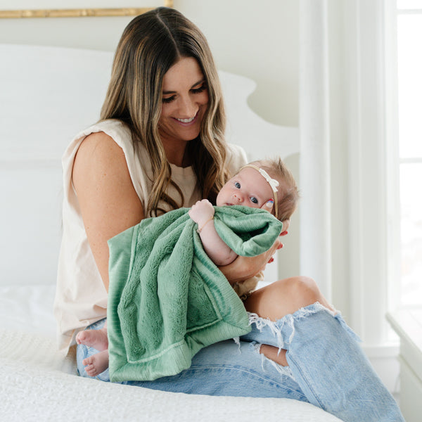 A mother holds her baby girl in a Mini Seagrass (dusty green) Colored Lush Saranoni Blanket. The soft blanket is a small blanket and a baby blanket or toddler blanket.