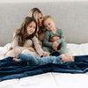 A mother and her daughter and two sons sit on a bed with a Toddler Navy Colored Lush Luxury Saranoni Blanket. The soft blanket is small blanket and a baby blanket or toddler blanket.