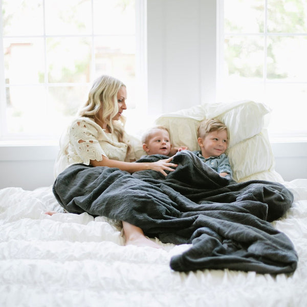 A mother and her two sons sit under a Luxury Extra Large Oversized Throw Charcoal Colored Lush Saranoni Blanket. The soft blanket is an extra large throw blanket and has a soft faux fur feeling.