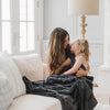 A mother and her daughter sit under a Luxury Toddler Charcoal Colored Lush Saranoni Blanket. The soft blanket is a toddler blanket or baby blanket.