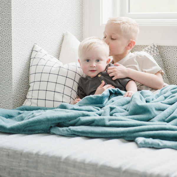 Two little boys sit under a Luxury Extra Large Oversized Throw Mineral Blue Colored Lush Saranoni Blanket. The soft blanket is an extra large throw blanket and has a soft faux fur feeling.