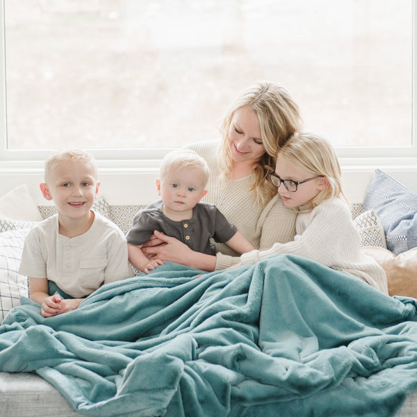 A mother sits with her two sons and daughter while holding a Luxury Extra Large Oversized Throw Mineral Blue Colored Lush Saranoni Blanket. The soft blanket is an extra large throw blanket and has a soft faux fur feeling.