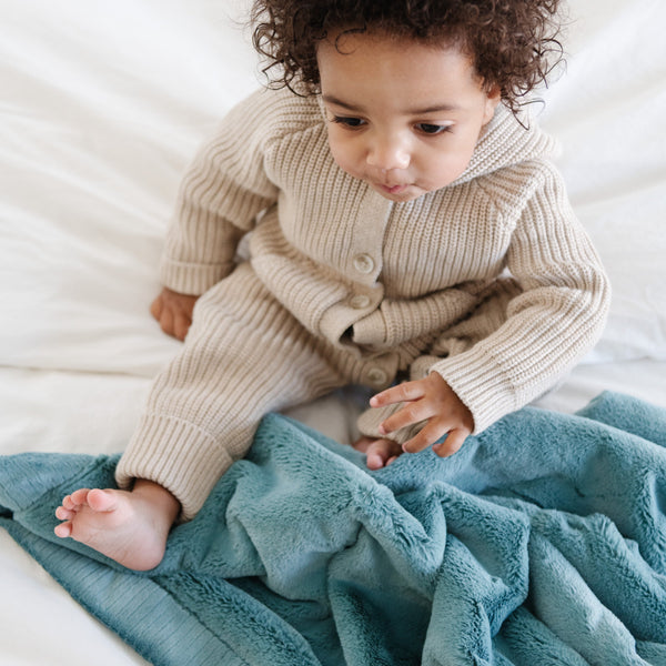 A little boy sits on a Luxury Receiving Mineral Blue Colored Lush Saranoni Blanket. The soft blanket is a small blanket and a baby blanket or toddler blanket.