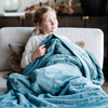 A mother sits with her two sonA little girl holds a Luxury Extra Large Oversized Throw Mineral Blue Colored Lush Saranoni Blanket. The soft blanket is an extra large throw blanket and has a soft faux fur feeling.