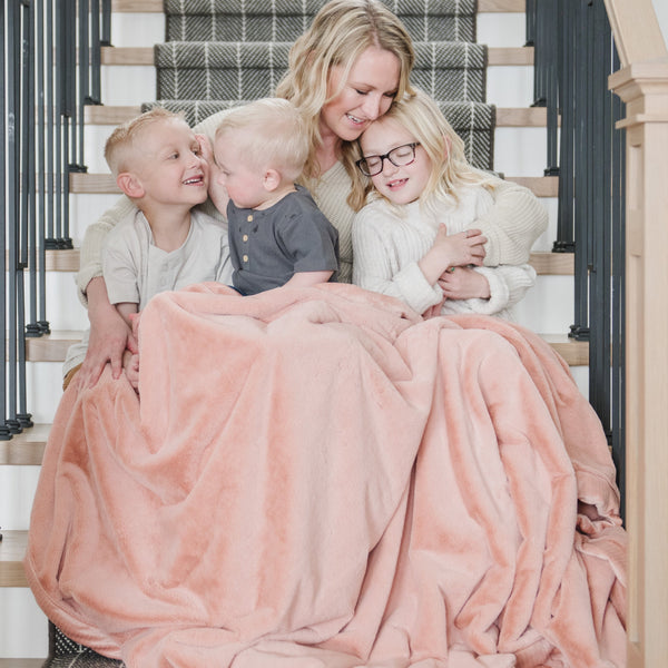 A mother and her daughter and two sons sit under a Luxury Extra Large Oversized Throw Blossom (light peachy pink) Colored Lush Saranoni Blanket. The soft blanket is an extra large throw blanket and has a soft faux fur feeling.