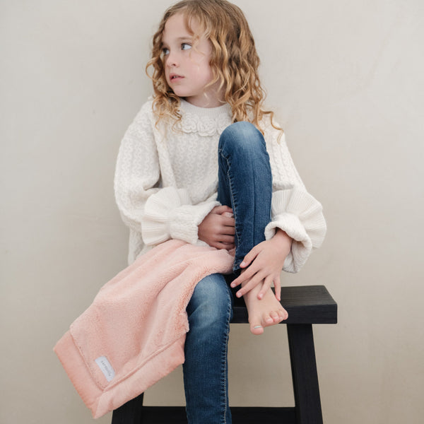 A little girl holds a Mini Blossom (light peachy pink) Colored Lush Saranoni Blanket. The soft blanket is a small blanket and a baby blanket or toddler blanket.