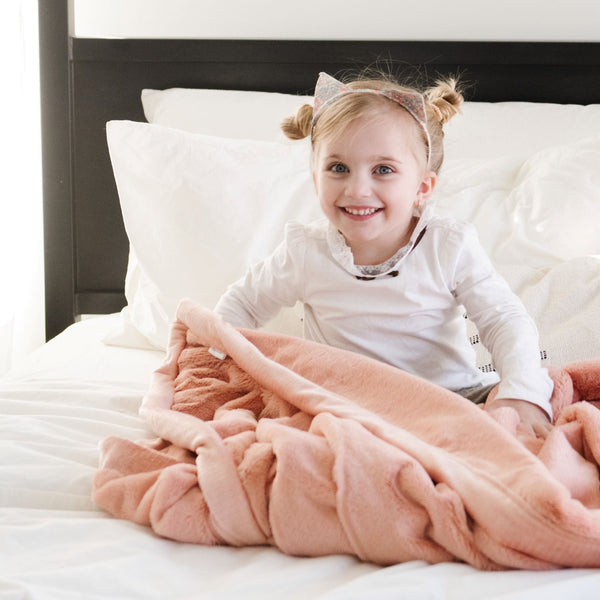 A little girl sits on a Blossom (light peachy pink) Colored Lush Saranoni Blanket. The soft blanket is a small blanket and a baby blanket or toddler blanket.