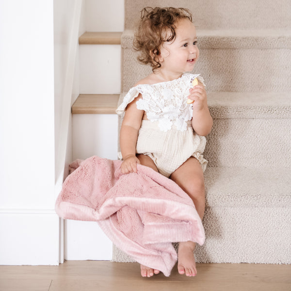 A little girl wearing a white outfit sitting on stairs holding a Mini Saranoni Lush Blanket that is light pink. The specific color is called Ballet Slipper.